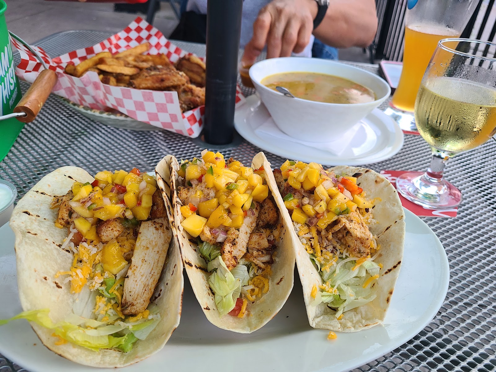 Three soft tacos with meat, lettuce, cheese and diced fruit, with other dishes and drinks on an outdoor table